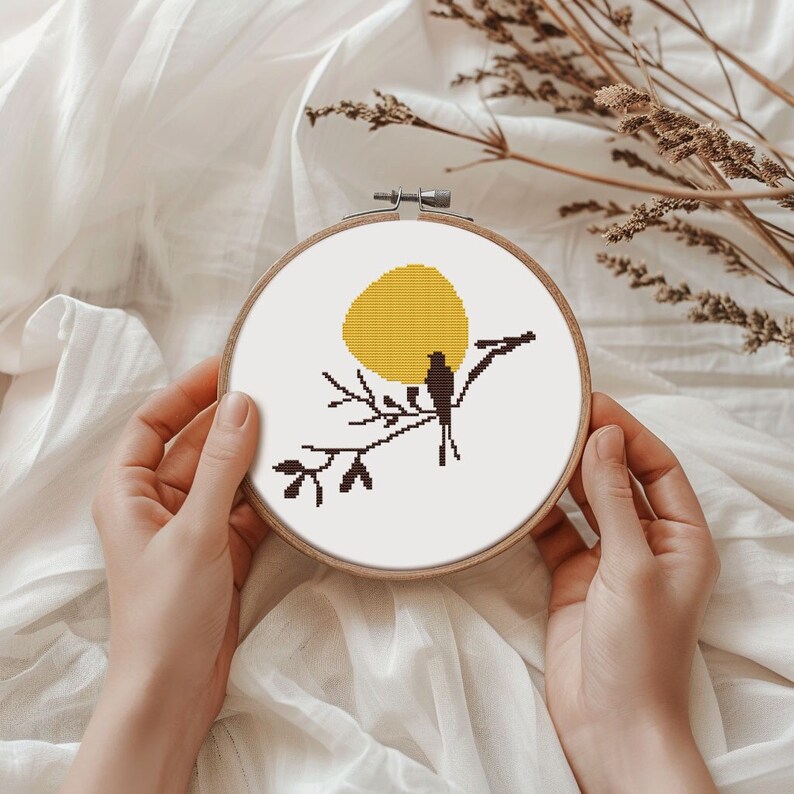 May include: A cross-stitch embroidery hoop with a minimalist design. The artwork shows a yellow sun, a bird on a branch, and black leaves. The embroidery is set in a wooden hoop, creating a rustic and artistic piece.