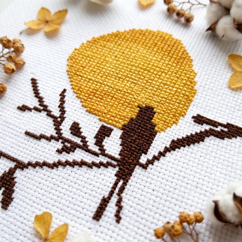 May include: A cross-stitch design depicting a brown bird on a branch, with a yellow sun against a white backdrop. The image is framed by dried flowers and cotton bolls, evoking a rustic, nature-inspired aesthetic.