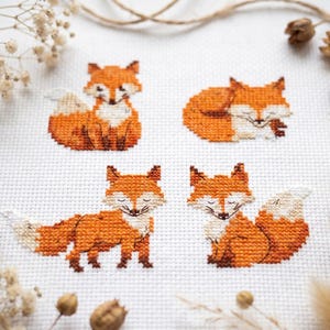 May include: A white fabric piece featuring four distinct cross-stitch patterns of orange and white foxes in various poses, including sitting, sleeping, and standing, rendered with detailed thread work on a grid-like textile surface.