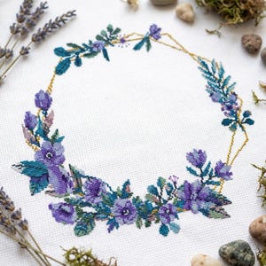 May include: A cross-stitch embroidery pattern on white fabric featuring a circular wreath of purple and blue flowers and green leaves, accented by a thin, geometric gold-colored hexagonal frame outline that encircles the floral arrangement.