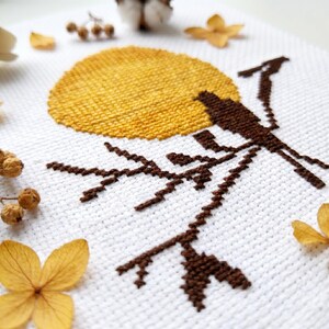 May include: A cross-stitch design showing a brown bird on a branch, with a yellow sun in the background. The embroidery is on white fabric, with dried flowers and seed pods around the edges. The design has a rustic, natural aesthetic.