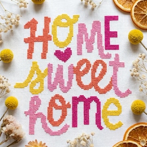 Peut inclure: Une broderie au point de croix sur tissu blanc avec le texte "HOME sweet HOME" en fil jaune, orange et rose. Un petit cœur rose est brodé entre les mots. Le motif est entouré de fleurs séchées et de tranches d'orange séchées.