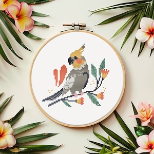 Puede incluir: Un bastidor de bordado de punto de cruz con una cacatúa colorida posada en una rama con flores. El ave tiene detalles grises, amarillos y negros. El bordado está sobre un fondo blanco, rodeado de hojas y flores tropicales.