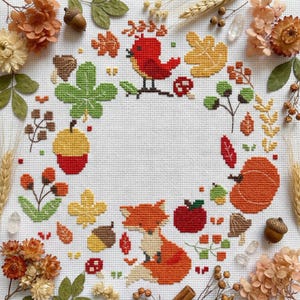 May include: A cross-stitch design featuring a red bird, fox, and various autumn elements like leaves, acorns, and a pumpkin. The design is set against a white background, with dried flowers and wheat surrounding the artwork.
