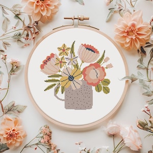 Puede incluir: Bordado de punto de cruz de un ramo floral en una taza gris con lunares, dentro de un bastidor de bordado de madera. Las flores son en tonos rosa, amarillo y blanco, con hojas verdes. El fondo es blanco, con flores color melocotón alrededor del bastidor.