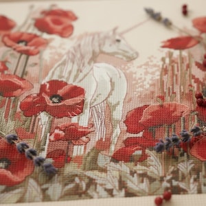 May include: Cross-stitch picture depicting a white unicorn amidst red poppies. The embroidery includes lavender and berries, creating a whimsical floral scene. The colour palette is soft, with shades of red, white, and green.
