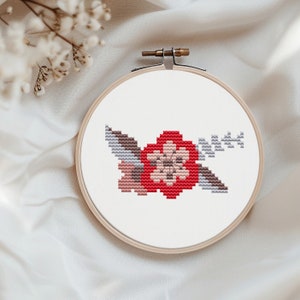 May include: A cross-stitch embroidery hoop with a floral design. The design features a red flower with pink and gray leaves.