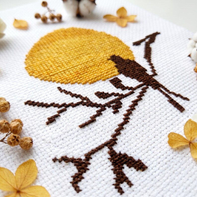 May include: Cross-stitch embroidery of a brown bird on a branch, with a yellow sun on a white backdrop. The design is framed by dried flowers and cotton buds, evoking a rustic, nature-inspired aesthetic. The piece is a handcrafted item.