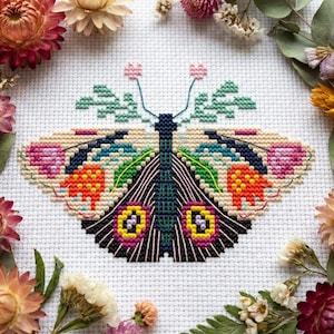 May include: A cross-stitch embroidery of a butterfly on white fabric. The butterfly features a dark blue body, wings with cream, orange, pink, and yellow patterns, and green leaf-like antennae topped with small pink flowers.
