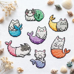 May include: Cross-stitch art featuring seven whimsical cat mermaids in various colors. Each cat has a different colored tail, including blue, purple, orange, and pink. The cats are surrounded by seashells and dried flowers on a white background.
