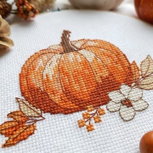 Può includere: Un ricamo a punto croce su tessuto bianco raffigurante una zucca arancione centrale con un picciolo marrone. La zucca è arricchita da un fiore bianco a cinque petali, piccole bacche arancioni e foglie dai colori autunnali.