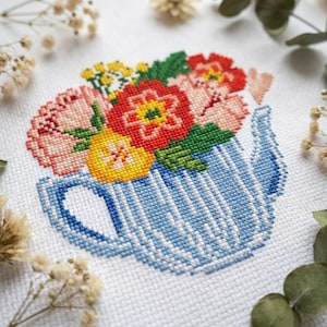 May include: A cross-stitch embroidery on white fabric featuring a blue and white striped teapot filled with a vibrant bouquet of red, yellow, and pink flowers, accompanied by a small stitched heart shape positioned to the right of the floral arrangement.