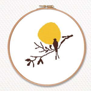 May include: A cross-stitch embroidery hoop with a yellow sun, a brown bird, and branches. The design is set against a white background. The embroidery is framed by a wooden hoop.