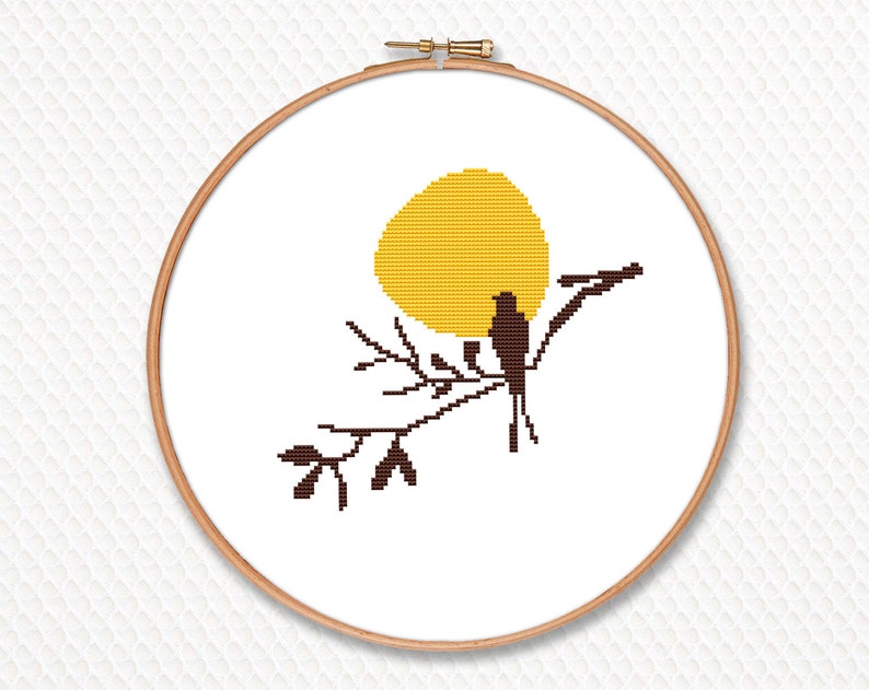 May include: A cross-stitch embroidery hoop with a yellow sun, a brown bird, and branches. The design is set against a white background. The embroidery is framed by a wooden hoop.
