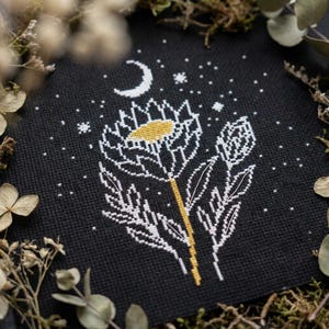 May include: A black fabric square featuring a white cross-stitch design of a large flower with a yellow center, a smaller flower bud, and a crescent moon surrounded by small stars. The embroidery is stitched directly onto the dark textile.