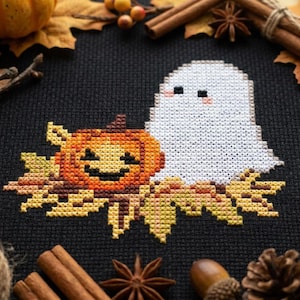 May include: A cross-stitch embroidery on black fabric featuring a white ghost with pink cheeks next to an orange jack-o'-lantern pumpkin. The design is surrounded by autumn-themed elements including brown maple leaves, cinnamon sticks, and acorns.