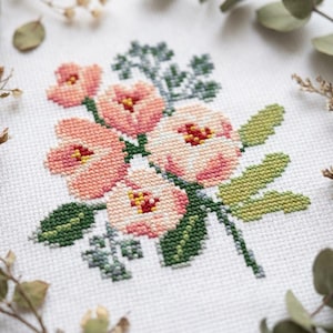 May include: A cross-stitch embroidery pattern on white fabric featuring a cluster of five flowers in shades of peach, pink, and cream with yellow centers, surrounded by green leaves and foliage stems in various shades of dark and light green.