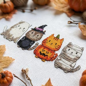 May include: A cross-stitch embroidery on white fabric featuring four cats in Halloween costumes: a ghost cat, a cat wearing a purple-tipped witch hat, a cat inside a jack-o'-lantern pumpkin, and a cat wrapped in white bandages like a mummy.
