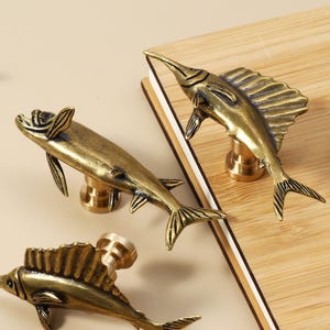 May include: Four antique brass fish-shaped cabinet pulls. The pulls are detailed with fins, tails, and a long, pointed snout. They are arranged on a light-coloured wooden surface, suggesting a nautical or coastal theme.