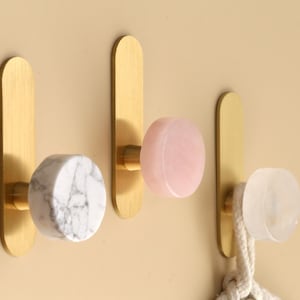 Könnte beinhalten: Drei dekorative Wandhaken mit messingfarbenen Rückplatten und runden Knöpfen. Die Knöpfe sind aus Marmor, rosa Stein und klarem Stein gefertigt. Ein weißes Seil hängt an dem Haken aus klarem Stein. Die Haken sind an einer beigefarbenen Wand befestigt.