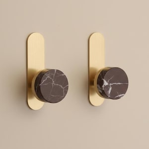 Op de afbeelding: Twee wandhaken met een goudkleurige, langwerpige achterplaat en een cirkelvormige, donkerbruine marmeren knop. Het marmer heeft witte aderen. De haken zijn op een neutraal gekleurde muur gemonteerd.