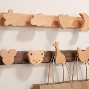 Könnte beinhalten: Zwei Wandgarderoben aus Holz mit dekorativen Formen. Die obere Garderobe hat einen Stern, eine Wolke, ein Herz und einen Mond. Die untere Garderobe zeigt eine Maus, einen Bären, eine Giraffe und eine Katze. Die Garderoben sind aus hellem und dunklem Holz gefertigt.