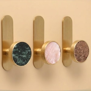 May include: Three wall hooks with brass-coloured backplates and circular knobs. The knobs have green, pink, and brown stone-like textures. The hooks are mounted on a light beige wall.