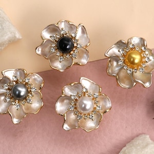 May include: Four flower-shaped decorative knobs with pearl centers and rhinestone accents. The knobs have a gold-colored trim and are set against a pink and beige background. The center pearls are black, gold, gray, and white.