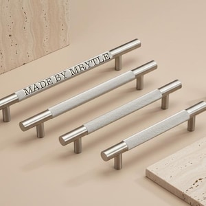 Op de afbeelding: Een set zilverkleurige kastgrepen en een knop. De grepen hebben een getextureerd, lichtgekleurd middengedeelte en zijn voorzien van het opschrift "MADE BY MYRTLE". De hardware is geplaatst op een neutrale achtergrond.