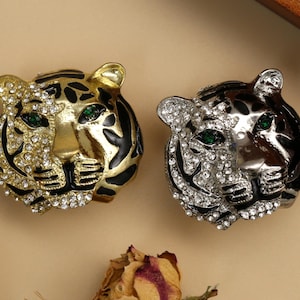 May include: Two decorative tiger head brooches. One is gold-toned with black stripes and clear rhinestones, the other is silver-toned with black stripes and clear rhinestones. Both have green gem eyes.