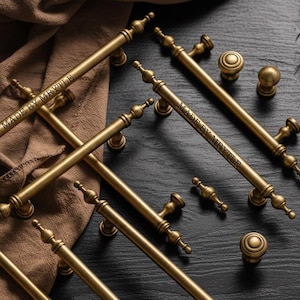 Puede incluir: Una colección de herrajes de latón antiguo para armarios, que incluye tiradores largos, pomos y tiradores decorativos más pequeños. El texto "MADE BY MRVTLE" es visible en los tiradores. Los herrajes están dispuestos sobre una superficie oscura.