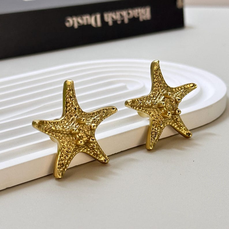 K&ouml;nnte beinhalten: Ein Paar goldfarbene Seestern-Ohrringe. Die Ohrringe sind mit strukturierten Oberfl&auml;chen und kleinen, erhabenen Noppen versehen. Die Ohrringe werden auf einer wei&szlig;en, geschwungenen Oberfl&auml;che pr&auml;sentiert.