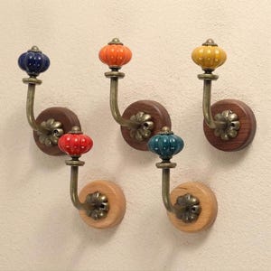 May include: Five wall-mounted hooks featuring brass-toned metal arms and round wooden backplates. Each hook is topped with a decorative, ribbed ceramic knob in a different colour: dark blue, bright orange, yellow, red, and teal green.