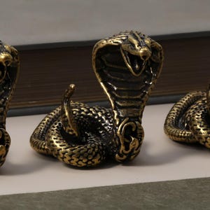 Pode incluir: Close-up de várias figuras de cobra de bronze. Cada cobra está enrolada com o capuz levantado, revelando detalhes intrincados de escamas e bocas abertas. As figuras têm um acabamento metálico dourado e bronze escuro.