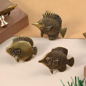 May include: Bronze-coloured fish-shaped cabinet knobs and pulls. The knobs and pulls are in various sizes and designs, including a long fish pull and several round fish knobs. The knobs and pulls are made of metal and have a detailed, textured finish.