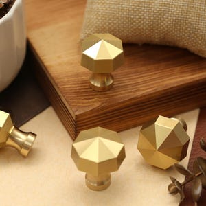 May include: Four gold-colored metal cabinet knobs with a faceted, multi-sided geometric top and a smooth, tapered base. The knobs feature a polished finish that reflects light, creating distinct highlights and shadows on their angular surfaces.