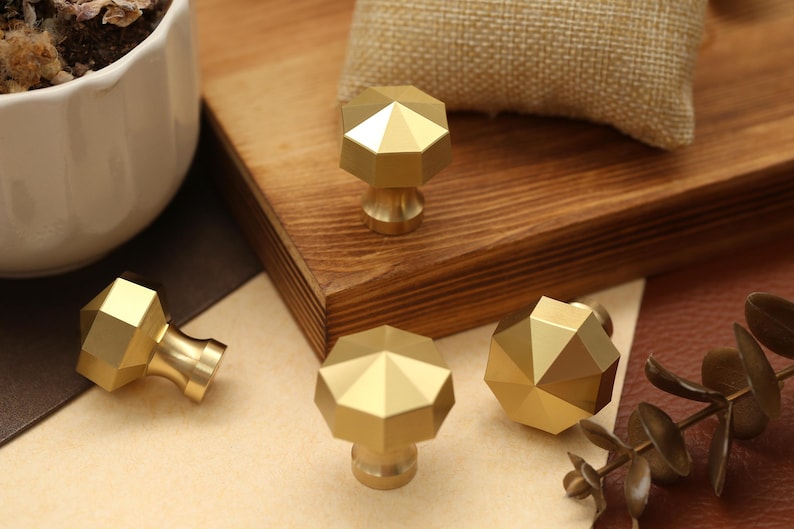 May include: Four gold-colored metal cabinet knobs with a faceted, multi-sided geometric top and a smooth, tapered base. The knobs feature a polished finish that reflects light, creating distinct highlights and shadows on their angular surfaces.