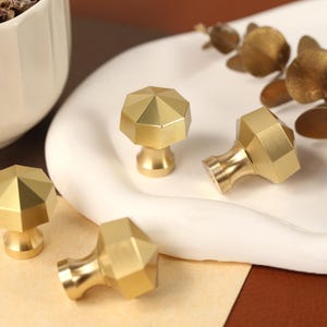 May include: Four brass cabinet knobs with a faceted, octagonal top surface and a smooth, tapered base. Each knob features a metallic gold finish with a matte texture, showcasing a geometric design that reflects light across its angled edges.