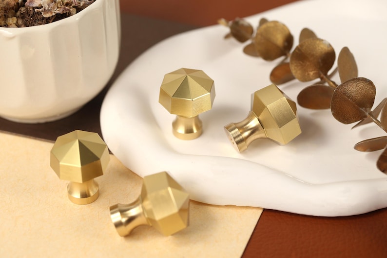May include: Four brass cabinet knobs with a faceted, octagonal top surface and a smooth, tapered base. Each knob features a metallic gold finish with a matte texture, showcasing a geometric design that reflects light across its angled edges.