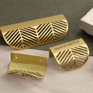 May include: Gold-coloured cabinet pulls with a leaf-like design. The pulls have a semi-circular shape with a cut-out pattern. One pull has the text "MADE BY MYRTLE". The pulls are made of metal and are on a neutral background.