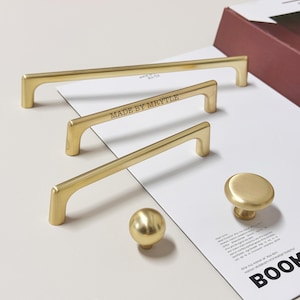 Puede incluir: Una colección de herrajes dorados para armarios, que incluye tres tiradores de barra, un pomo redondo y un pomo esférico. El tirador central lleva grabado "MADE BY MRYTLE". Los herrajes están dispuestos sobre una superficie blanca con papel impreso.