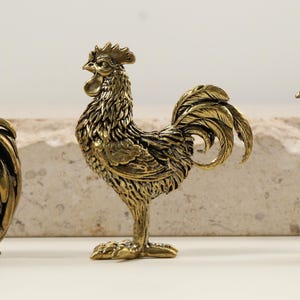 Puede incluir: Tres detalladas figuras de gallos de latón. El gallo central se alza erguido, con sus plumas y cresta intrincadamente esculpidas. Los otros dos gallos también son de latón, con detalles precisos. Las figuras se colocan sobre un fondo neutro.
