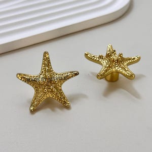 May include: Two gold-coloured starfish cabinet knobs. The knobs have textured surfaces and are in a five-point star shape. The text "MADE BY MRYTLE" is visible on one of the knobs. These knobs are suitable for furniture or decor.