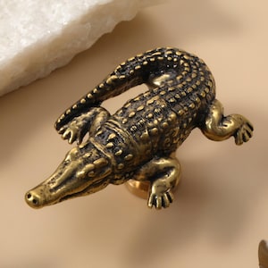 Op de afbeelding: Een gedetailleerde messing kastknop in de vorm van een alligator. De knop heeft een goud-bronzen kleur, met getextureerde schubben en een gedetailleerde snuit en poten. De alligator is geplaatst op een lichtbeige oppervlak, met een witte steen op de achtergrond. Een uniek decoratief hardware-onderdeel.