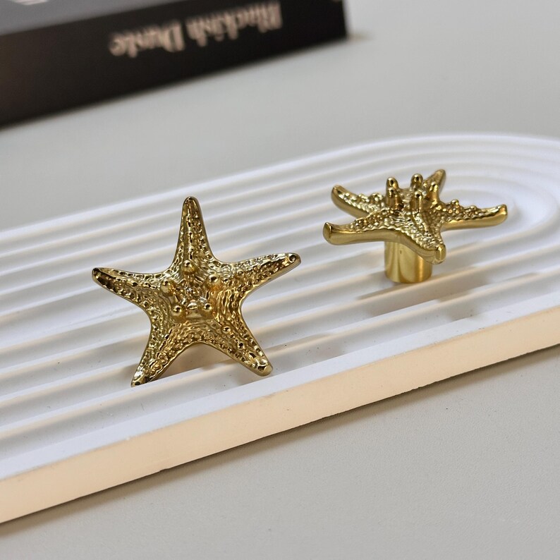 K&ouml;nnte beinhalten: Zwei goldfarbene, sternf&ouml;rmige Schrankkn&ouml;pfe. Die Kn&ouml;pfe sind aus Metall und haben eine detaillierte, strukturierte Oberfl&auml;che. Ein Knopf liegt flach, w&auml;hrend der andere auf einem kurzen Pfosten steht. Die Kn&ouml;pfe befinden sich auf einem wei&szlig;en, gebogenen Tablett.