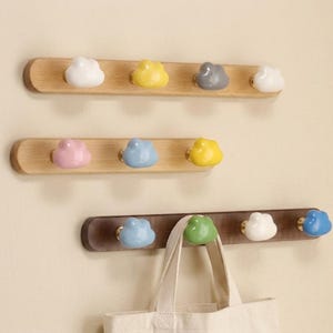 May include: Three horizontal wall-mounted racks made of wood, featuring rounded edges and multiple ceramic hooks shaped like clouds in various colors including white, yellow, gray, pink, blue, and green, with a canvas bag hanging from one hook.