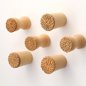 May include: A set of six cylindrical wall hooks made of light-toned wood. Each hook features a flat, circular front face inlaid with a woven rattan pattern in a natural tan color. The hooks have a smooth, polished finish and a uniform design.