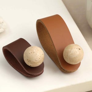 May include: Two leather drawer pulls in dark brown and tan, each featuring a smooth, rounded, beige stone knob attached to the center. The leather strips are folded into a loop shape, showcasing a natural texture and visible grain on the surface.