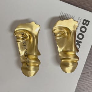 Könnte beinhalten: Zwei goldfarbene Ohrringe in Gesichtsform. Die Ohrringe haben eine strukturierte Oberfläche und sind im Profil. Der Text "MADE BY MRYTLE" ist vertikal auf dem linken Ohrring aufgedruckt. Auf einer weißen Oberfläche.