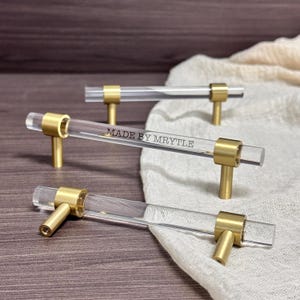 May include: Clear acrylic cabinet pulls with gold-coloured hardware. The pulls have a rectangular shape and are labelled "MADE BY MRYTLE". The hardware is cylindrical and connects the acrylic bar to the cabinet.